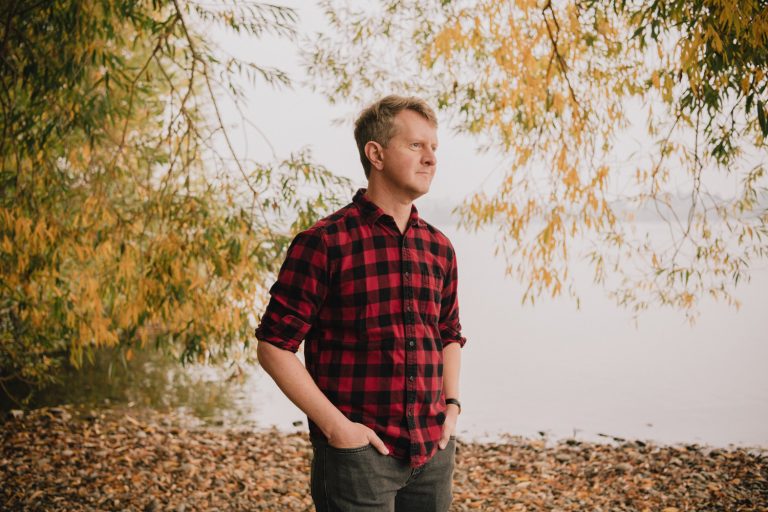 Adult male in a red and black plaid shirt stands with hands in pockets near water and golden autumn trees on an overcast day.