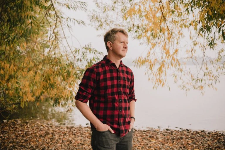 Adult male in a red and black plaid shirt stands with hands in pockets near water and golden autumn trees on an overcast day.