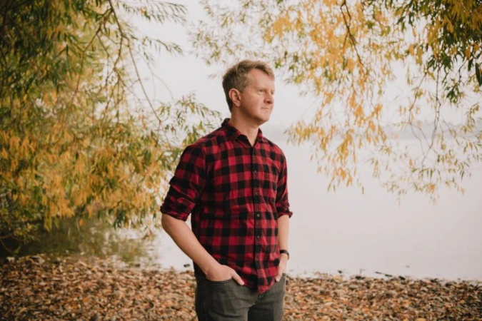 Adult male in a red and black plaid shirt stands with hands in pockets near water and golden autumn trees on an overcast day.