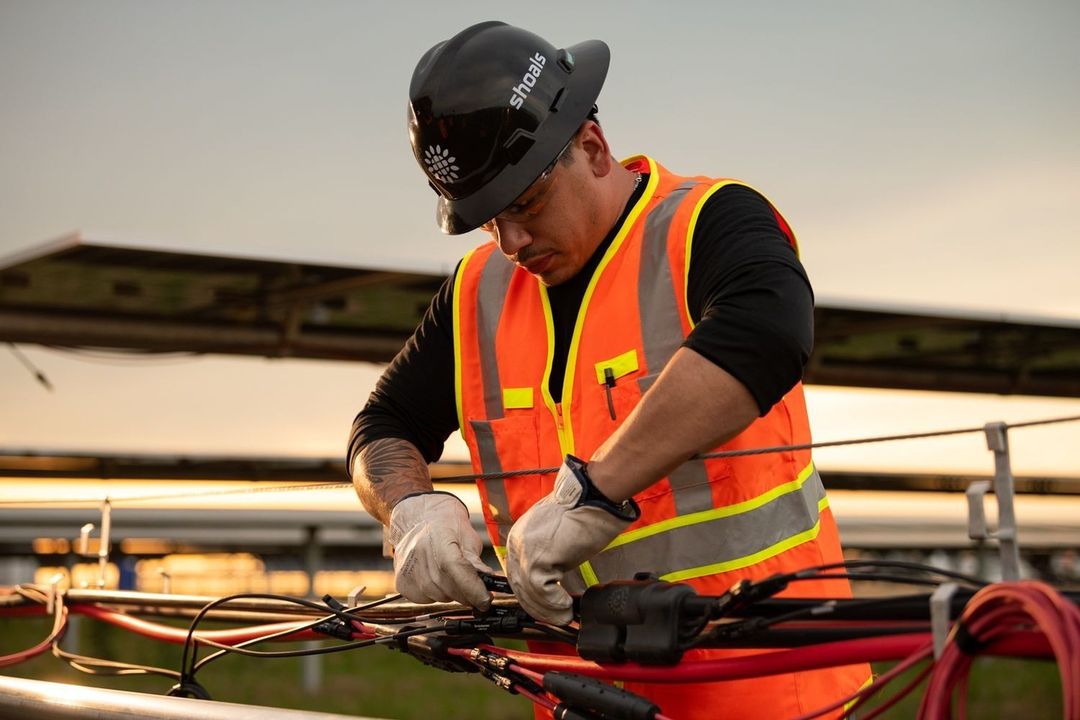 Man in safety gear working on solar panel wiring. He wears a Shoals hard hat and reflective vest in an outdoor environment.