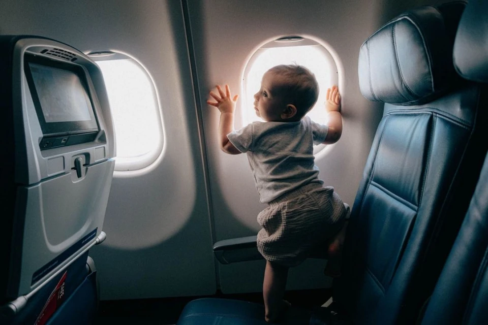 Baby boy stands on airplane seat looking out the window during a flight, showcasing travel and family.