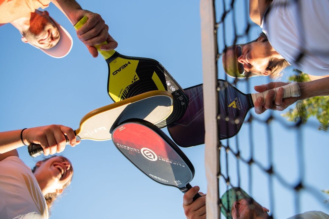 A group of pickleball players holding their rackets up in the air in a circle, showing unity and teamwork against a bright blue sky.