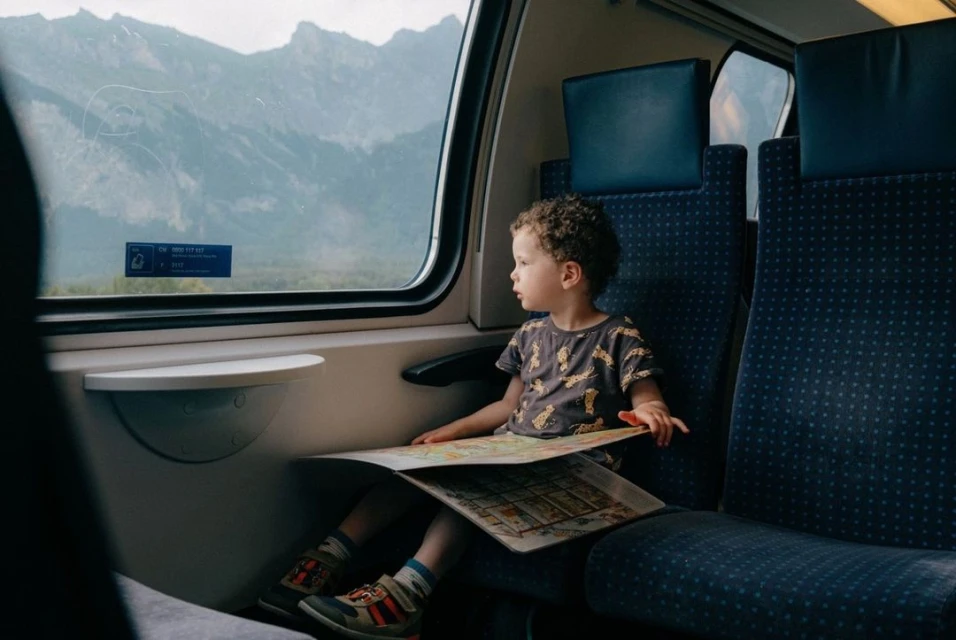 A child sits by a train window looking out at a mountain landscape, holding a map, with blue patterned seating.