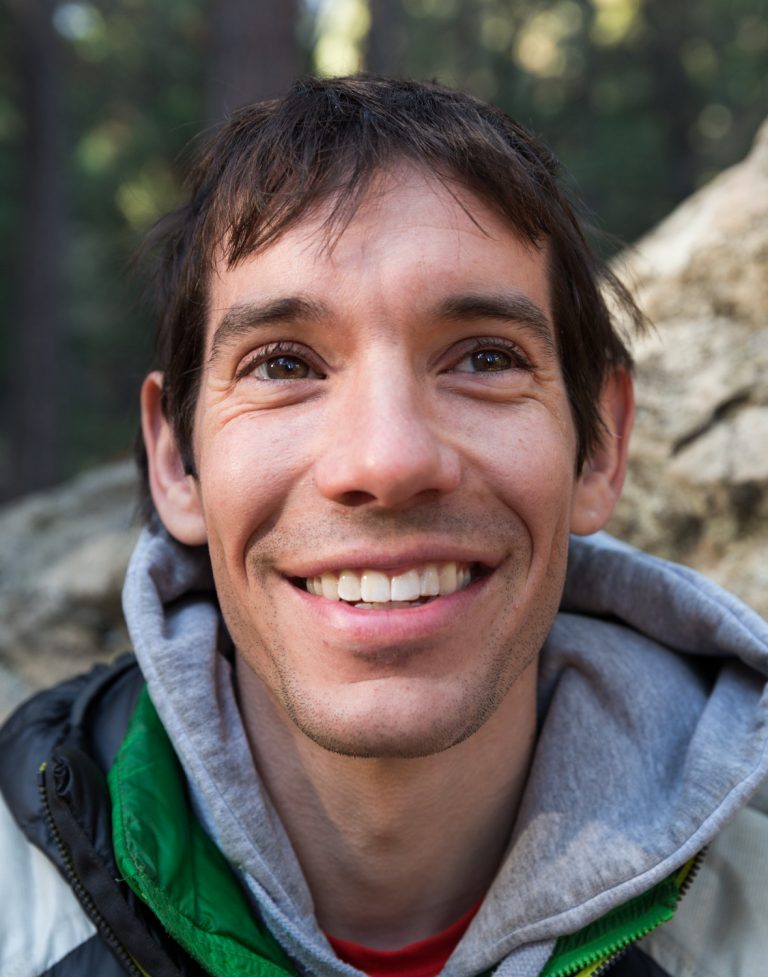 Portrait of Alex Honnold, smiling outdoors, wearing a grey hooded sweatshirt and green jacket, with a blurry rock and forest background.