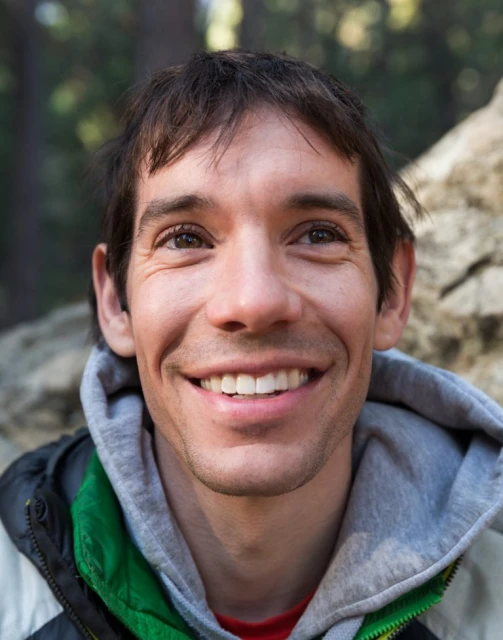 Portrait of Alex Honnold, smiling outdoors, wearing a grey hooded sweatshirt and green jacket, with a blurry rock and forest background.