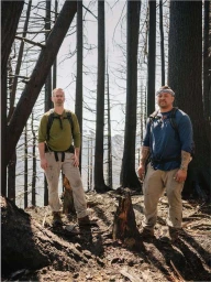 Two men standing in a burnt forest wearing hiking gear and backpacks amidst charred tree trunks