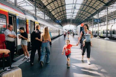 Mother holding child's hand walking on a busy railway station platform beside passenger trains