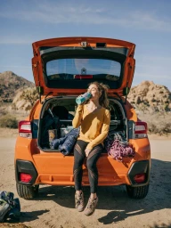 Woman sitting in the open trunk of an orange car drinking from a bottle outdoors in a desert landscape