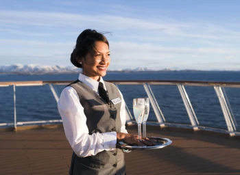 Smiling female steward in grey vest holding a silver tray with two champagne glasses on a cruise ship deck.