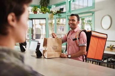 Delivery courier with orange backpack handing paper bag to customer at counter