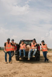 Five construction workers in hard hats and high-visibility vests posing by a black pickup truck on a dirt site.