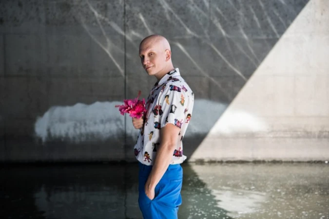 Bald man wearing a patterned short-sleeve shirt and blue trousers holding pink flowers near a concrete wall