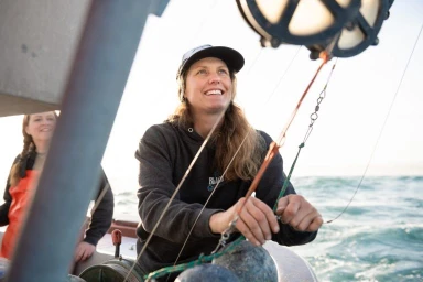 Smiling woman wearing a dark hooded sweatshirt and cap on a boat deck, with ocean water visible in the background.