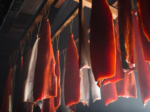 Multiple strips of orange salmon fillets hanging from wooden rafters in a smoky interior