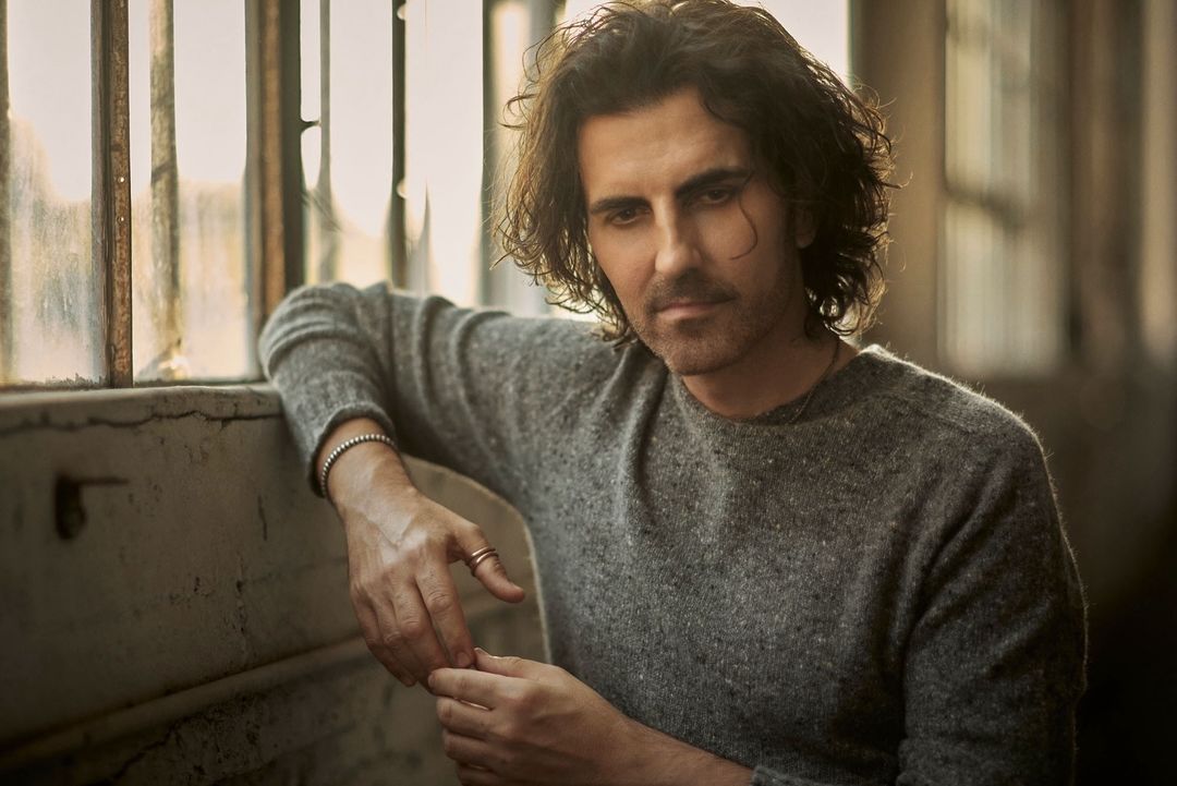Indoor portrait of an adult male with dark curly hair wearing a grey sweater leaning against a window with neutral-toned, soft lighting.