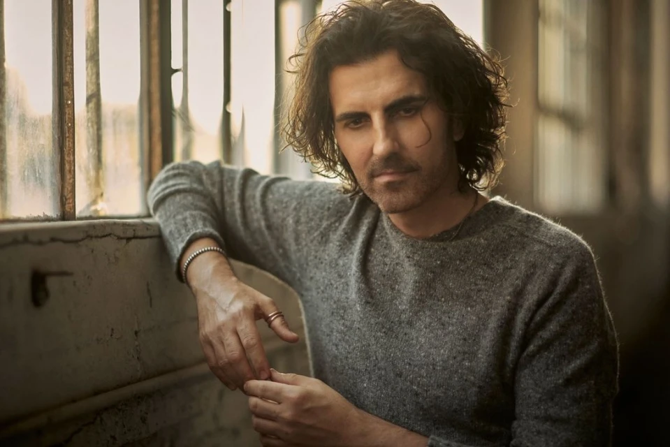 Indoor portrait of an adult male with dark curly hair wearing a grey sweater leaning against a window with neutral-toned, soft lighting.