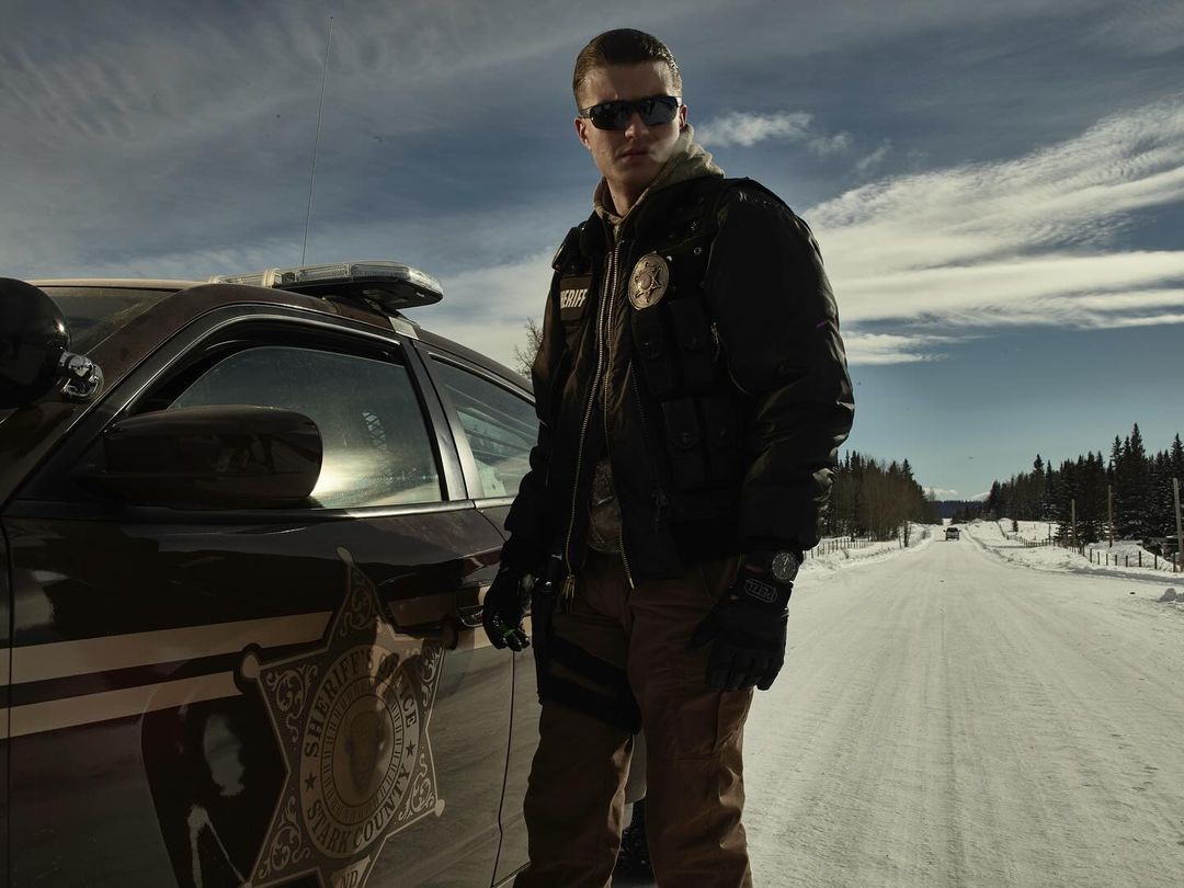 Adult male police officer stands near patrol car on snowy road under cloudy sky. Vehicle from Stark County Sheriff's Department.