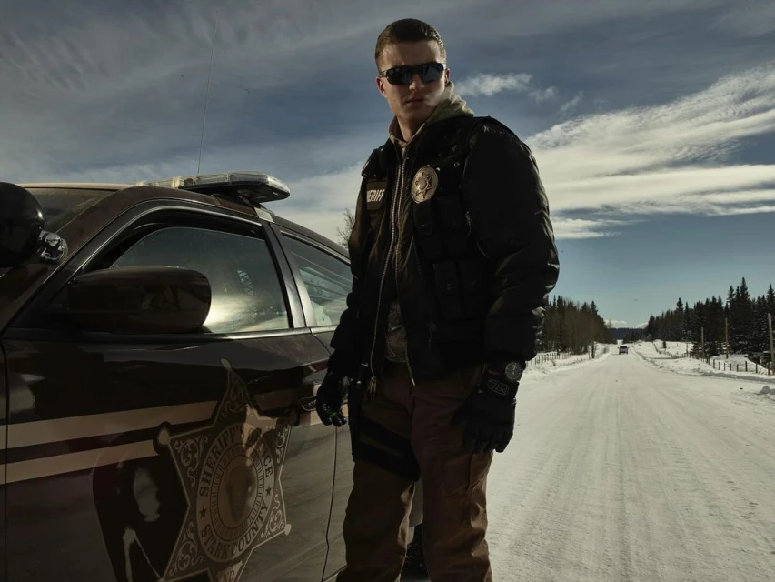 Adult male police officer stands near patrol car on snowy road under cloudy sky. Vehicle from Stark County Sheriff's Department.