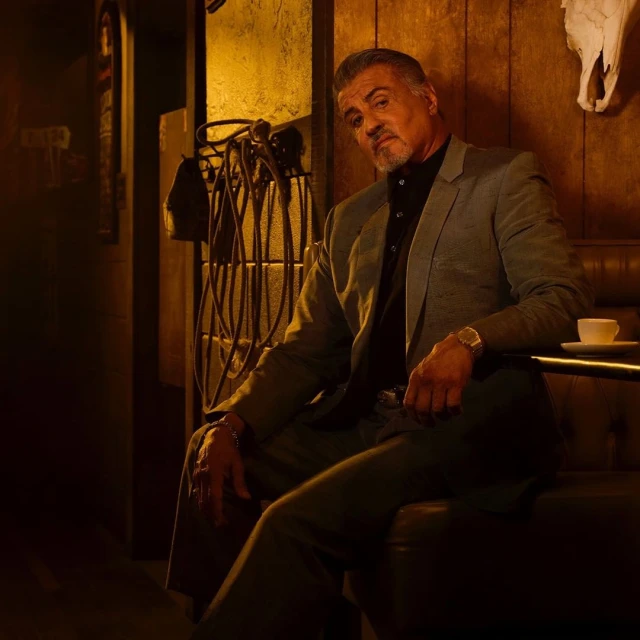 Portrait of an adult male in a suit jacket sitting indoors, with a cup on the table and an animal skull on the wall in the background.