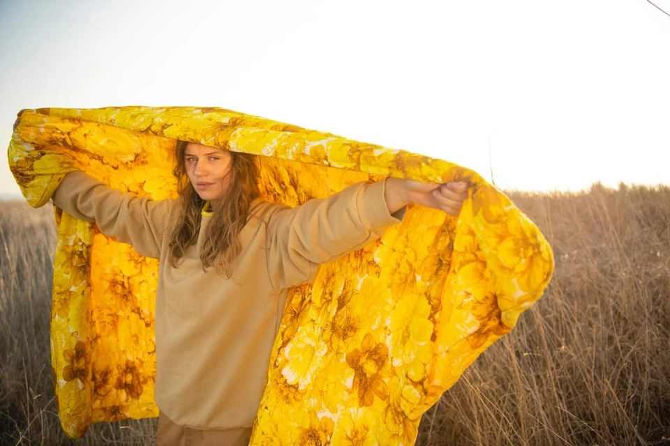 A young woman stands in a field holding up a large, bright yellow floral patterned textile as a light breeze blows through the dry grass.