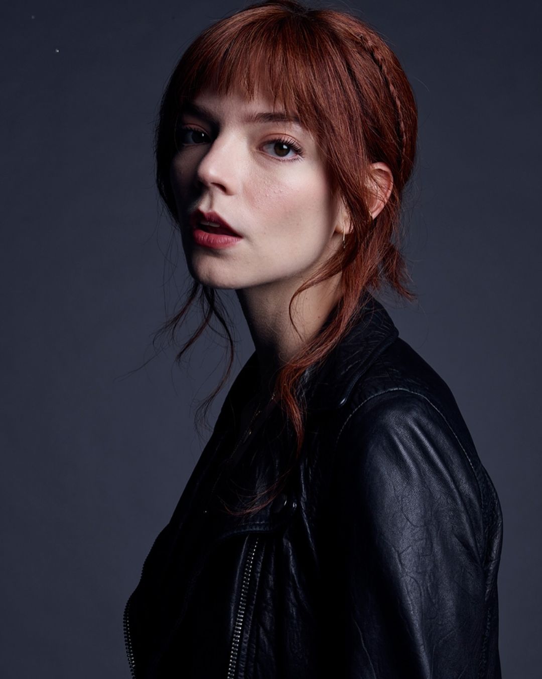 Portrait of a woman with red hair and a black leather jacket against a grey background, looking directly at the camera.