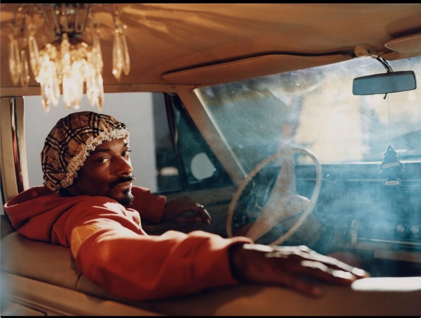 Adult male seated in a car wearing a bonnet and a red hoodie, looking towards the camera with a chandelier hanging above.