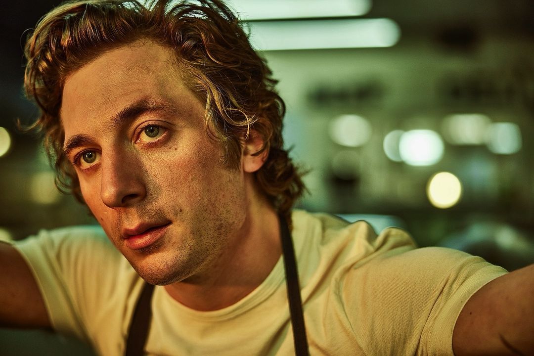 Close-up shot of a blonde adult male wearing a white t-shirt and black apron, looking up toward camera with a slight smile indoors.