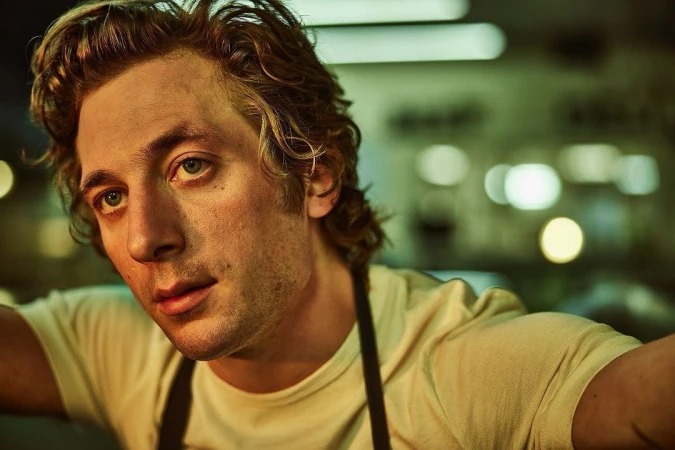 Close-up shot of a blonde adult male wearing a white t-shirt and black apron, looking up toward camera with a slight smile indoors.