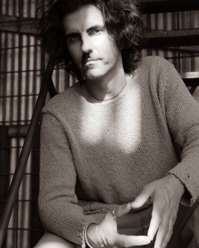 Monochrome portrait of a man with curly hair, wearing a sweater, sitting on metal stairs, bathed in sunlight creating shadows.