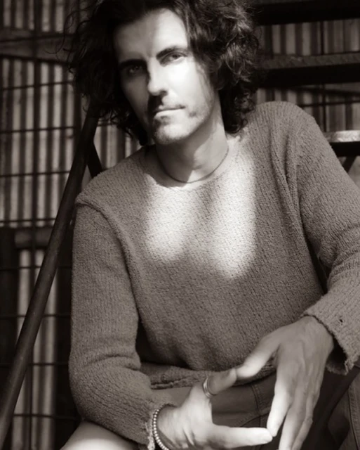 Monochrome portrait of a man with curly hair, wearing a sweater, sitting on metal stairs, bathed in sunlight creating shadows.