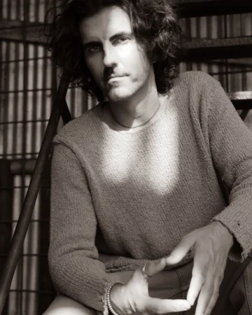 Monochrome portrait of a man with curly hair, wearing a sweater, sitting on metal stairs, bathed in sunlight creating shadows.