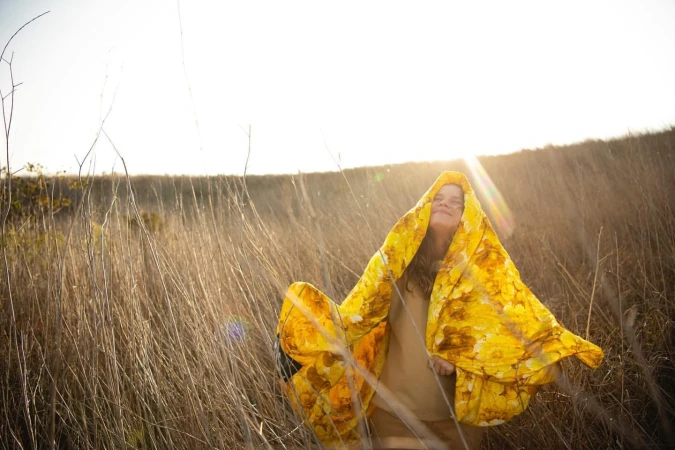 Woman wrapped in a bright yellow floral blanket standing in a tall dry grass field outdoors with sunlight