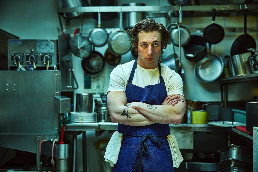 Close-up of a man in a chef's apron standing in a commercial kitchen with pots, pans, and stainless steel appliances in the background.