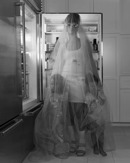 Black and white portrait of an adult woman in a kitchen with two dolls, wearing a veil, standing in front of an open refrigerator.