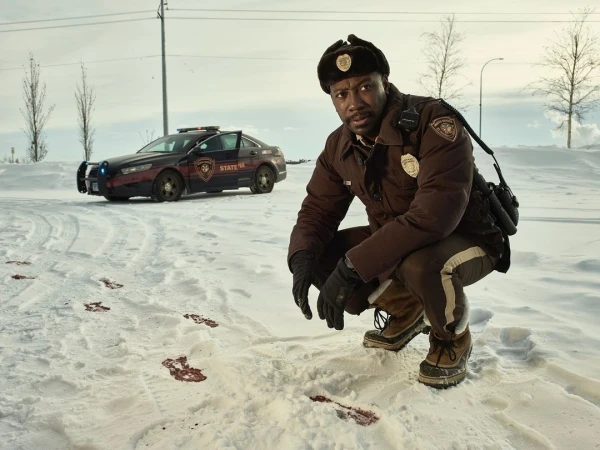 An African-American police officer in winter gear kneels in the snow, examining bloody footprints, with a patrol car in the background.