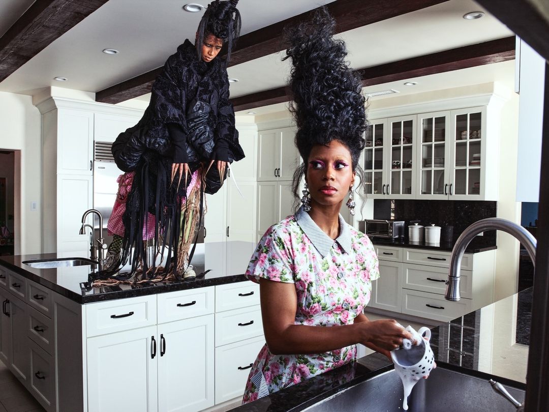 Two women in a kitchen, one washing dishes with an exaggerated hairstyle and the other atop the kitchen island wearing avant-garde fashion.