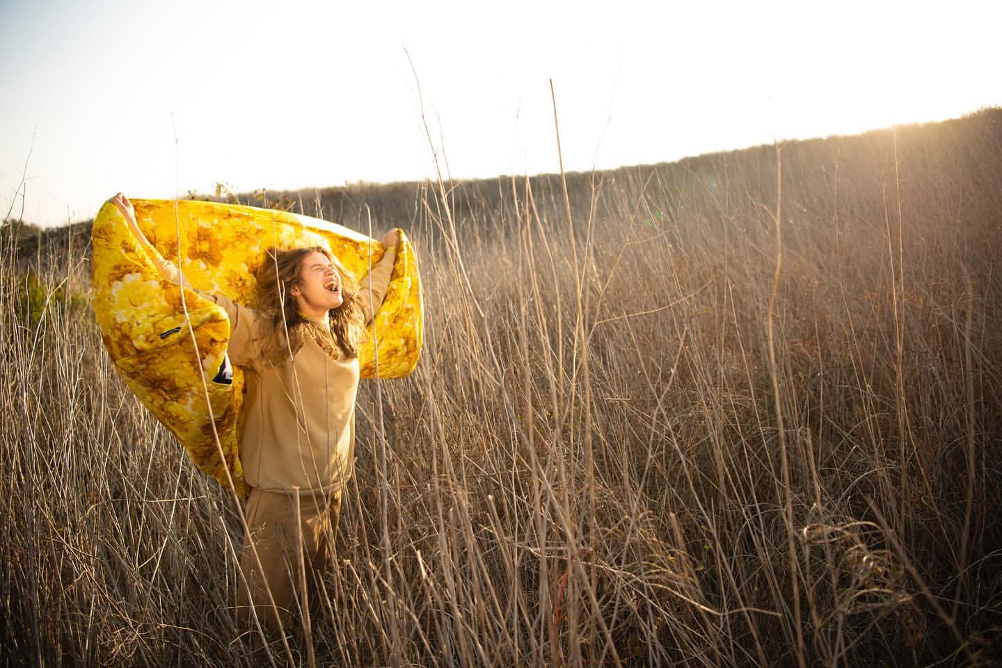 Woman in field holds up yellow patterned fabric, her face expressing joy in the outdoor setting during golden hour.