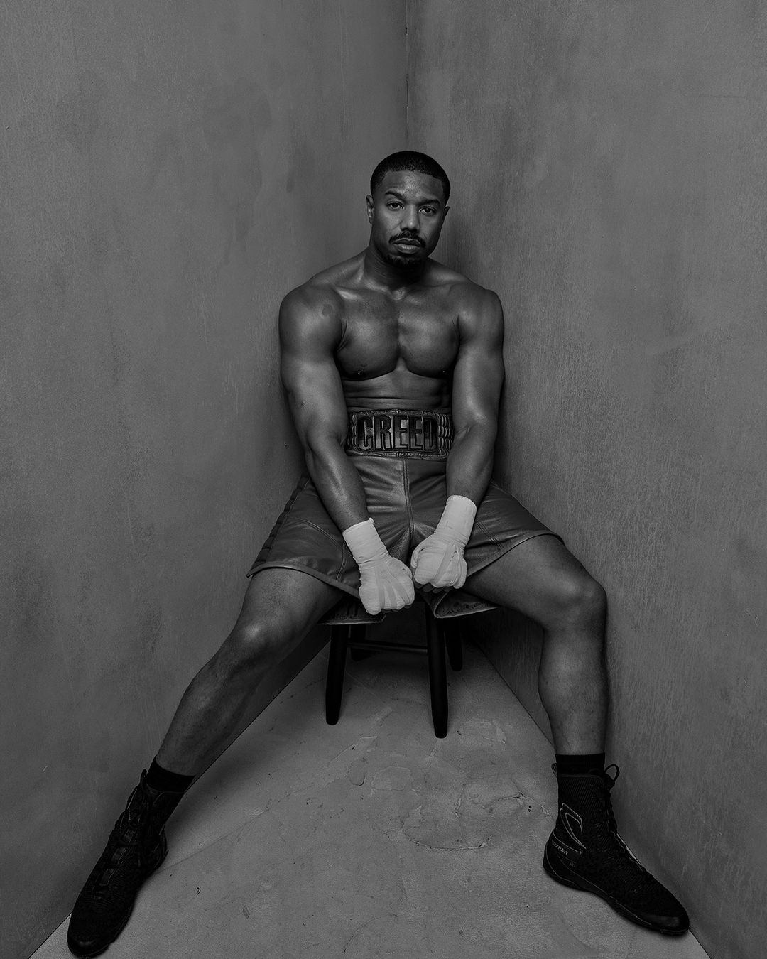 Black and white portrait of muscular adult male athlete sitting on a chair in boxing shorts and boots in an indoor corner setting.
