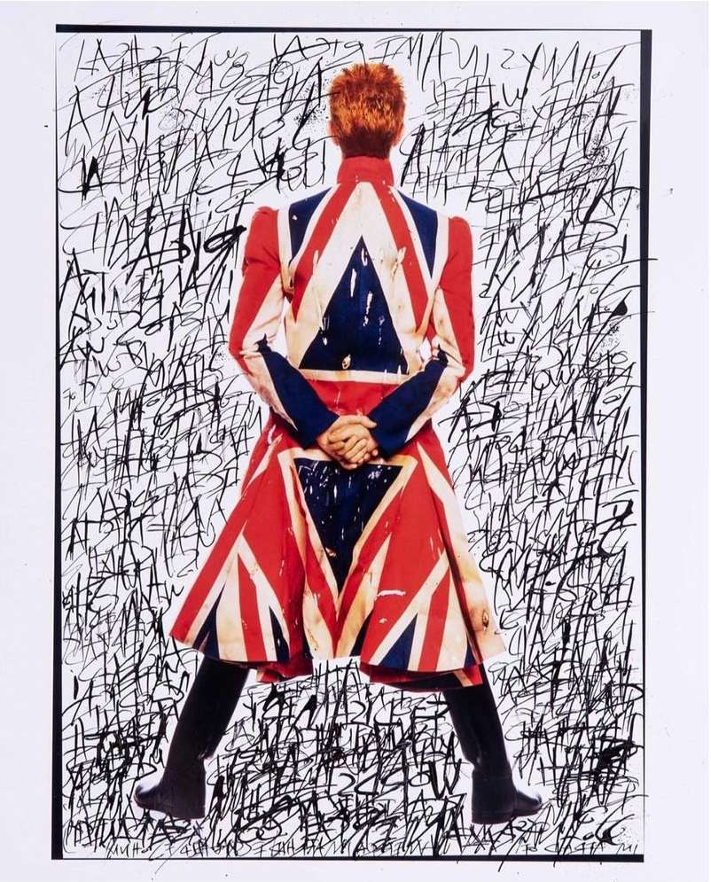 Back view of a person with orange hair wearing a Union Jack coat against a backdrop of dense black ink scribbles on white.
