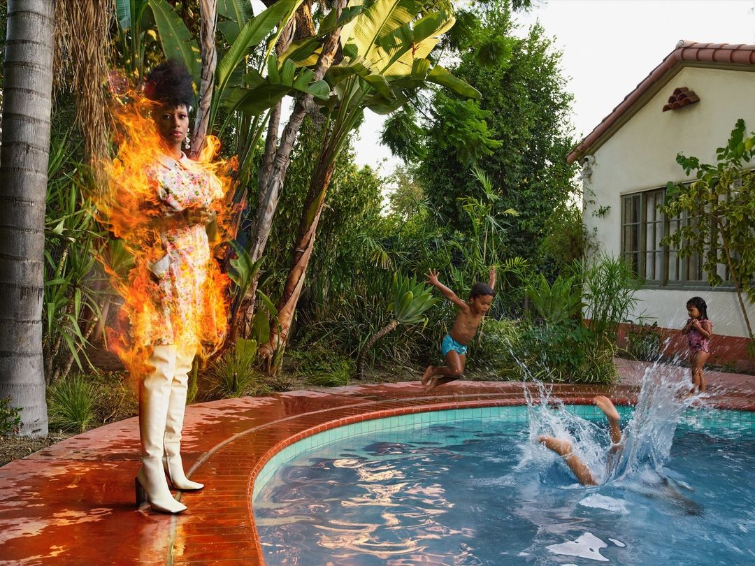 Woman in floral dress surrounded by flames poses near pool as children jump in, set against lush green tropical background.