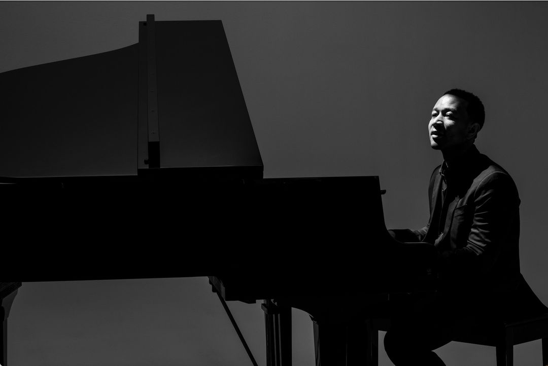 Black and white photo of a man sitting at a grand piano, bathed in light, with a plain background, showcasing musical performance.