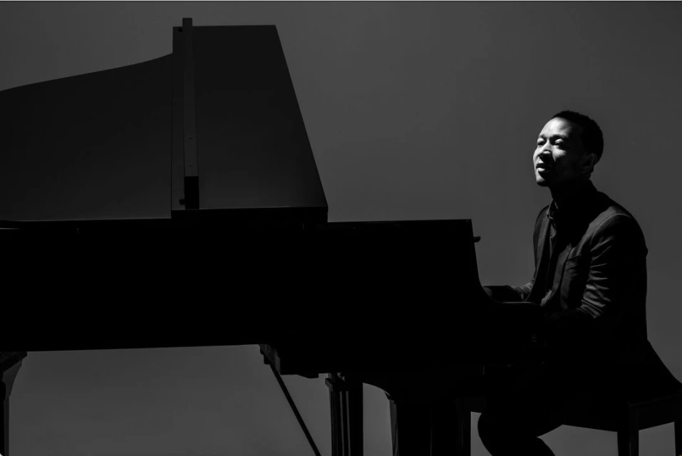 Black and white photo of a man sitting at a grand piano, bathed in light, with a plain background, showcasing musical performance.