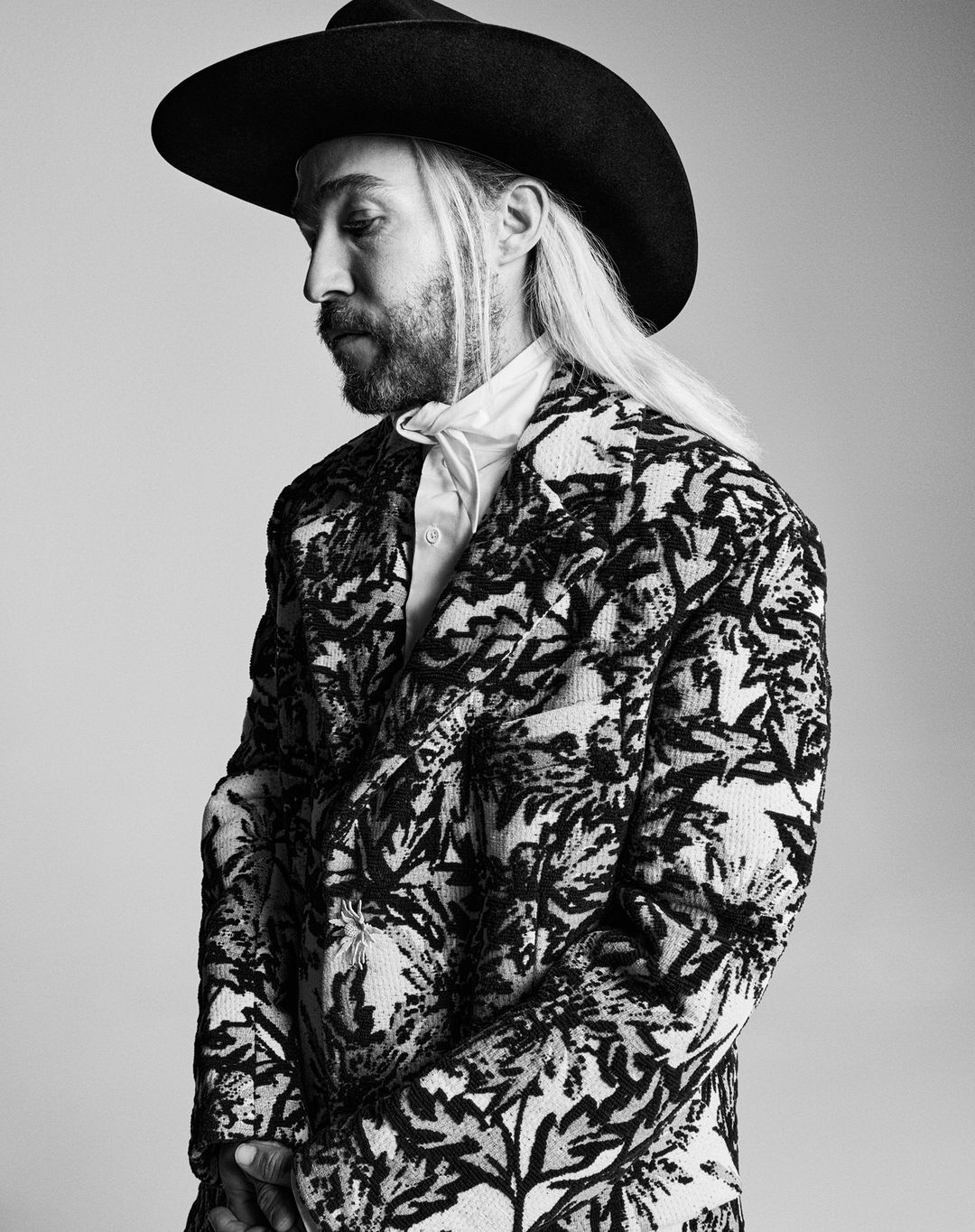 Black and white portrait of a fair-skinned adult male with a beard and long hair wearing a cowboy hat and patterned jacket.