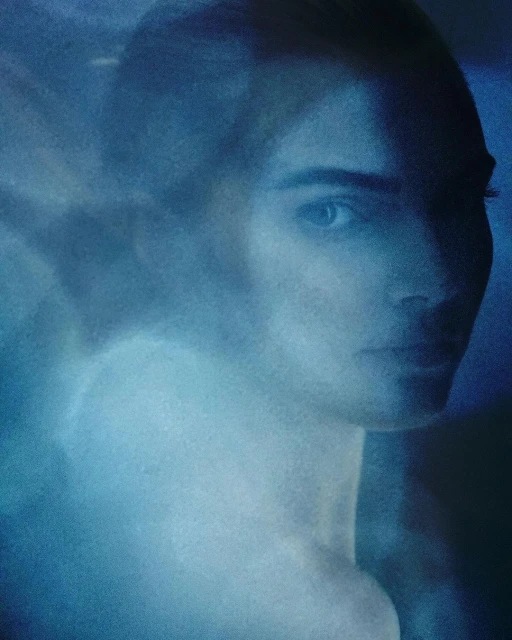 Close-up shot of an adult woman with neutral facial expression in an indoor setting with diffused lighting and a blue color grade.