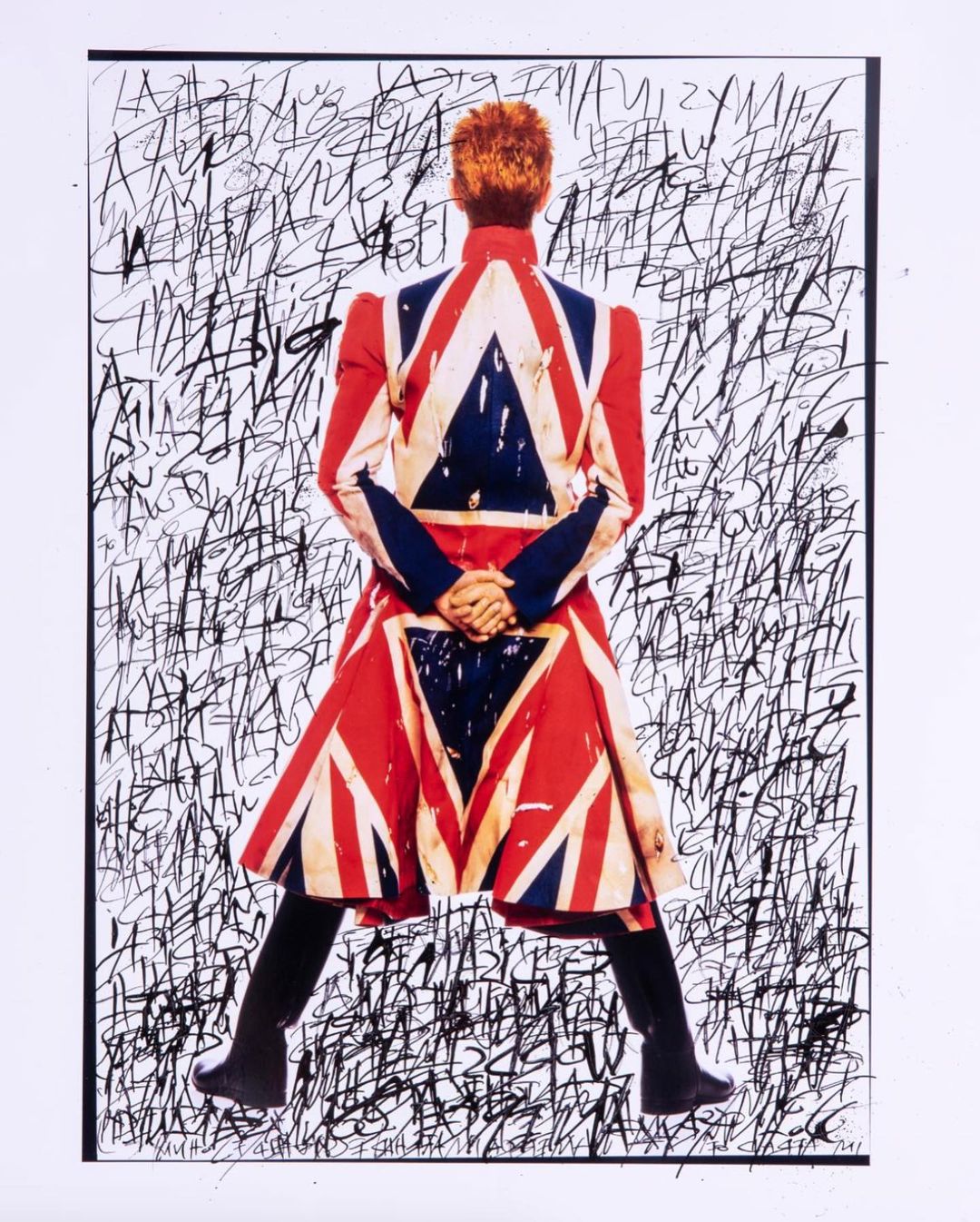 David Bowie, from behind, wears a jacket printed with the British Union Jack flag, against a backdrop of black scribbled text.