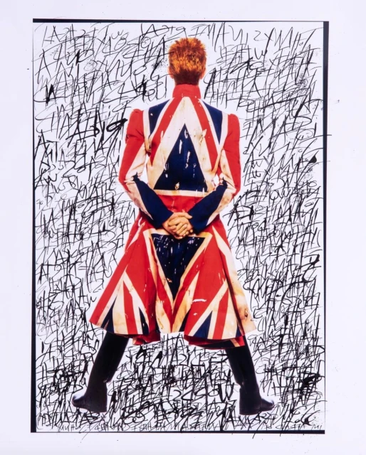David Bowie, from behind, wears a jacket printed with the British Union Jack flag, against a backdrop of black scribbled text.