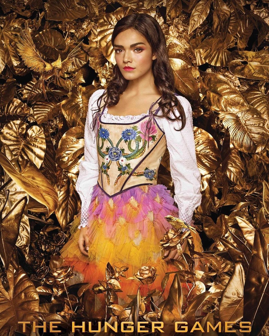 Portrait of female in decorative peasant blouse with feathery skirt and dark hair, posing against a gold floral background from 'The Hunger Games'.