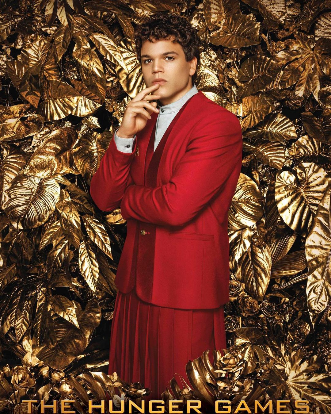 Portrait of adult male with curly hair, wearing a red suit and skirt, with hand to chin, posing in front of golden metallic foliage.