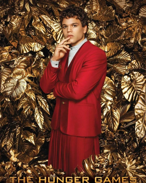 Portrait of adult male with curly hair, wearing a red suit and skirt, with hand to chin, posing in front of golden metallic foliage.