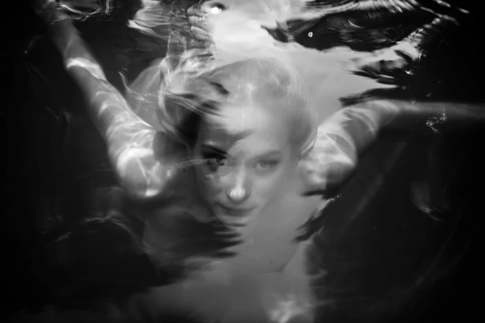 Black and white portrait of a woman submerged underwater, creating an ethereal, blurred effect with the water's reflections and refractions.